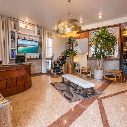 A stylish hotel lobby with a curved wooden reception desk, a TV wall, potted plants, gold pendant light, lounge seating, and a marble-tiled floor.
