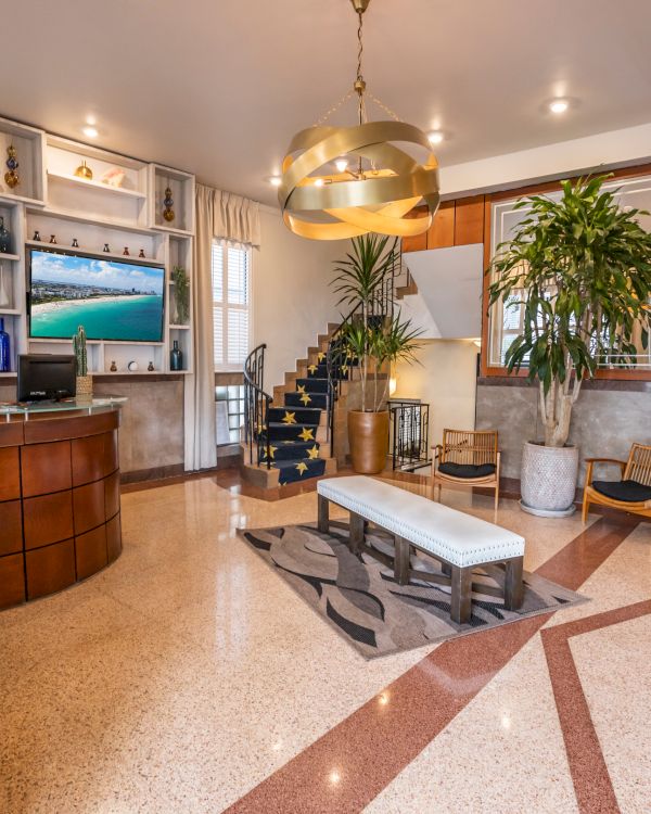 A stylish hotel lobby with a curved wooden reception desk, a TV wall, potted plants, gold pendant light, lounge seating, and a marble-tiled floor.