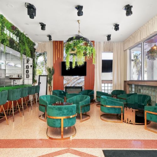 A stylish cafe interior with green seating, plants, a bar area, and warm lighting.