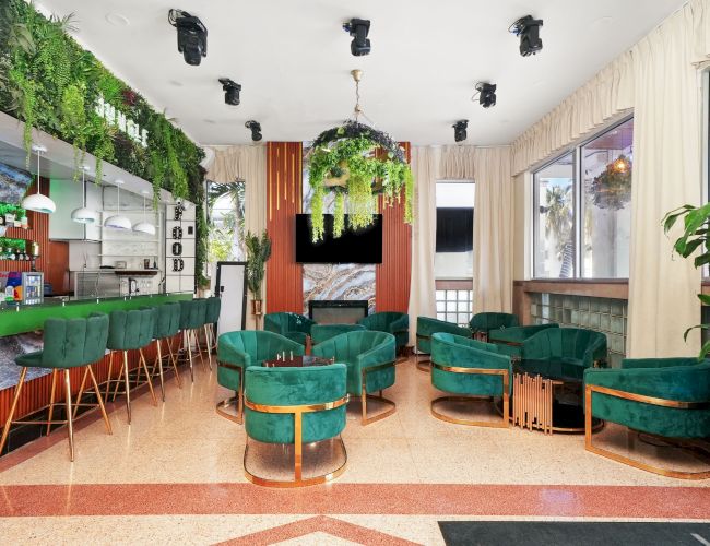 A stylish cafe interior with green seating, plants, a bar area, and warm lighting.
