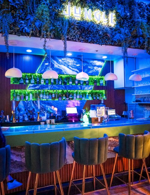 A neon-blue bar scene with hanging plants, blue lighting, and several green cushioned stools facing the counter, creating a stylish lounge vibe.