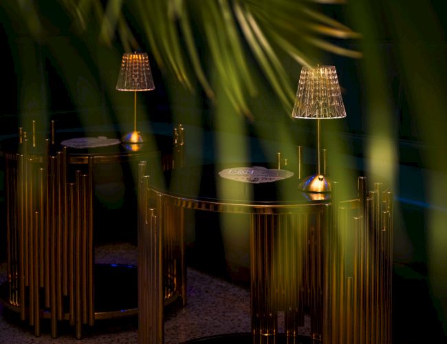 Dimly lit lounge with gold-accented chairs and hanging lamps, seen through plant fronds, inviting intimate nighttime seating.
