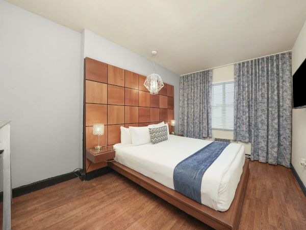 A modern hotel room with a wooden paneled headboard, queen bed, white linens, blue accents, wooden floor, desk, and window with patterned curtains.