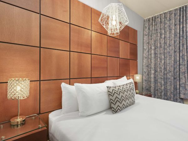 A modern hotel room with a padded wood headboard, white bedding, patterned throw pillow, nightstands, lamps, and a hanging pendant light.