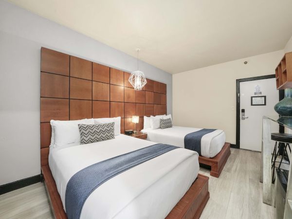 Two queen beds with white bedding and blue-gray runners, a wooden padded headboard, modern decor, and a small desk area in a bright hotel room.