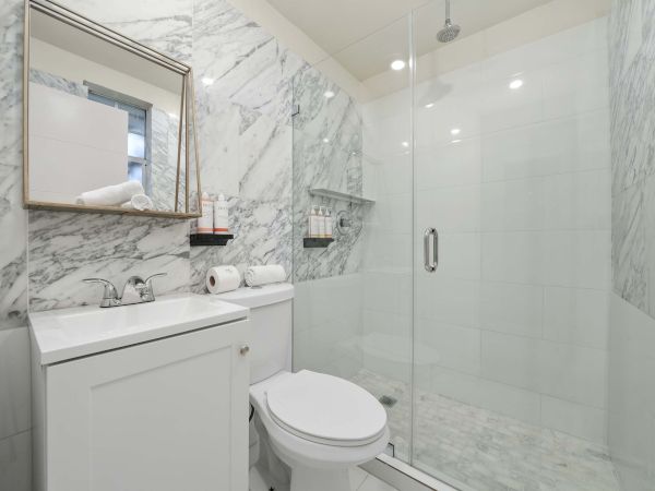 A modern marble bathroom with a glass shower enclosure, white vanity, toilet, and a rectangular mirror above the sink.