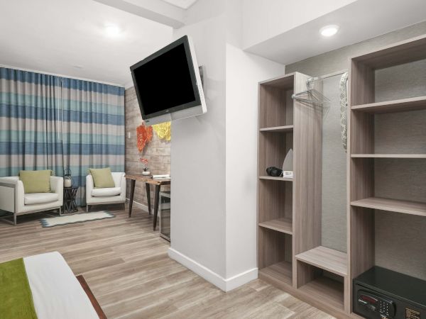 A modern hotel room with a seating area, wall-mounted TV, and open wardrobe/closet with shelves and a safe, plus a small desk and chair.
