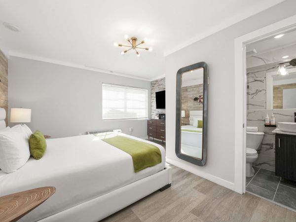 A bright hotel room with a white bed, green accents, a hanging mirror, and an open en-suite bathroom, airy and modern.