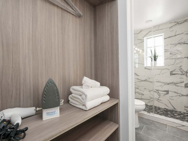 A small hotel closet with folded towels and a hairdryer on a shelf, next to a doorway leading to a marble-tiled bathroom.