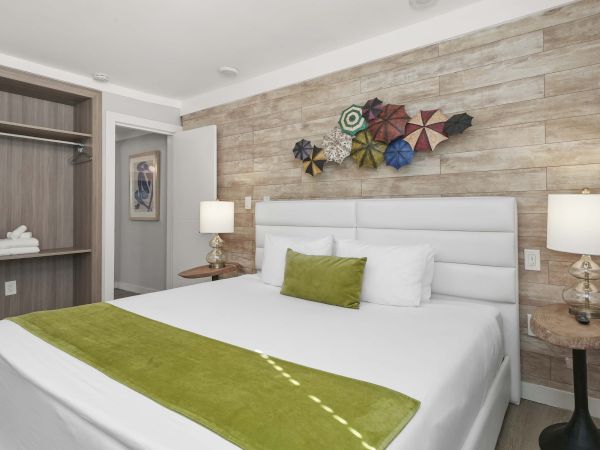 A neatly made hotel-style bedroom with a white bed, green accents, two lamps, a stone accent wall, and colorful wall art above the headboard.