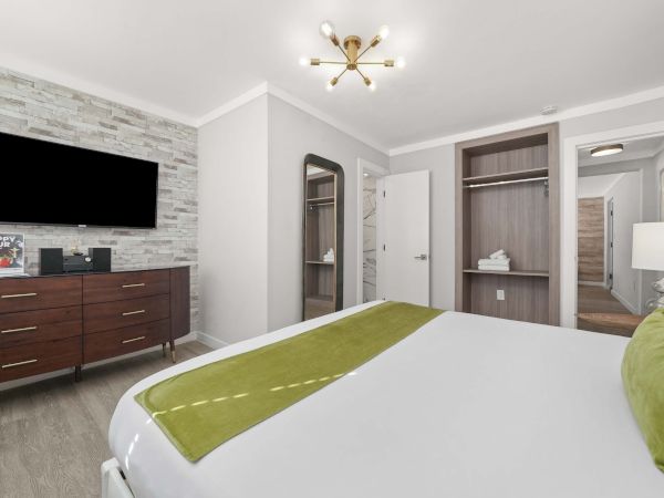 A modern hotel room with a large bed, green accents, wall-mounted TV, brick accent wall, dresser, and open wardrobe area by a doorway. End.