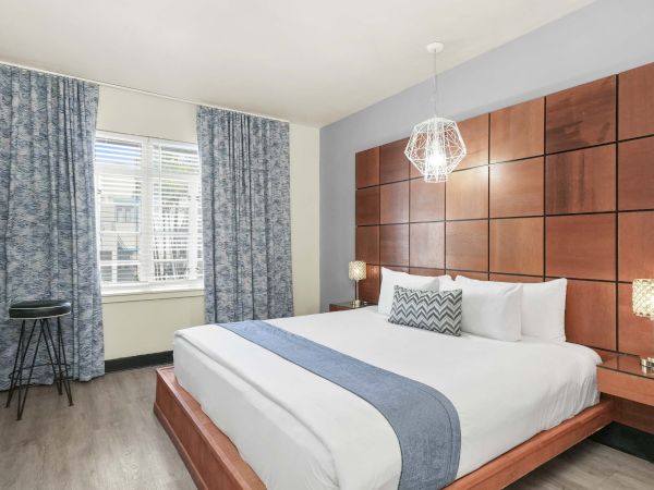 A modern hotel room with a large bed, wooden paneled headboard, white bedding with a blue runner, two nightstands, reading lamps, and blue curtains by a window.