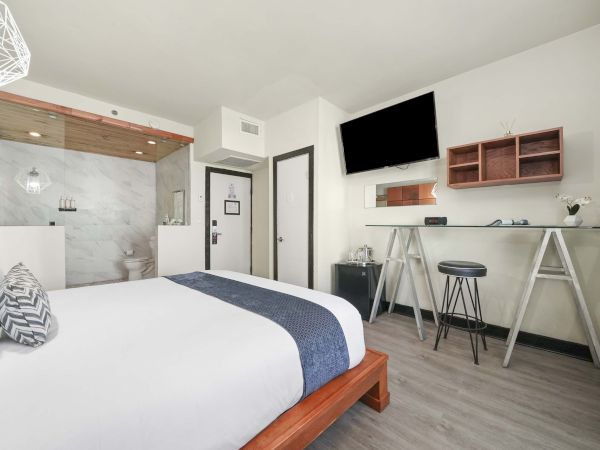 A cozy hotel room with a king bed, desk/sofa area, mounted TV, and an open-view kitchenette in a modern, minimal style.