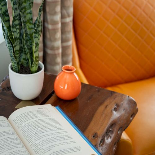 An open book on a rustic wooden table beside a white pot with a snake plant, an orange vase, and an orange quilted chair.