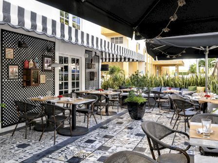 Outdoor cafe with black-and-white checkered wall, striped awnings, and many empty tables and chairs on a patterned tile terrace. End.