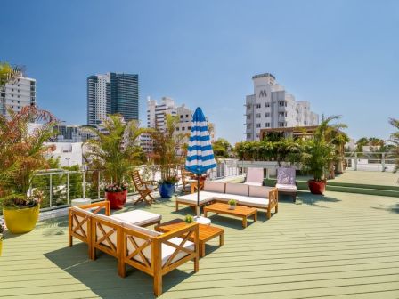 A sunny hotel rooftop terrace with wooden seating, potted plants, a blue striped umbrella, and city skyline in the background.