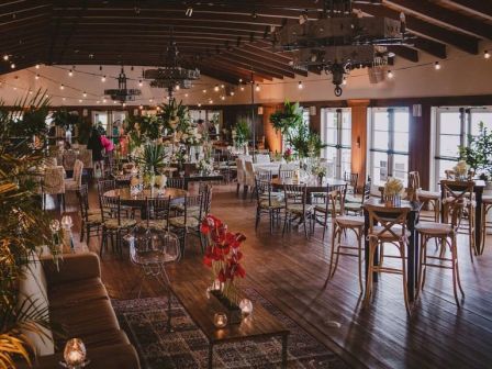 Stylish event venue with wooden floors, string lights, and high ceiling; chic tables, chairs, plants, and candles creating a cozy, elegant atmosphere.