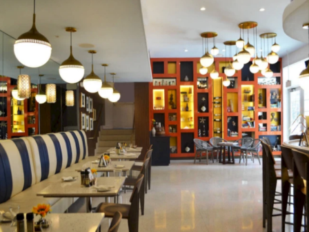 A stylish restaurant interior with blue-and-white booth seating, wooden tables set for dining, and warm pendant lights; orange display wall with shelves in the background.
