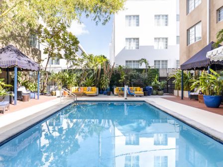 Pool surrounded by lounge chairs, palm trees, and shaded cabanas; blue water reflects a sunny courtyard with white buildings.