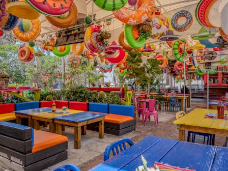A colorful outdoor seating area with rainbow cushions, bright tables, and hanging swim-ring decorations overhead, creating a fun, tropical vibe.