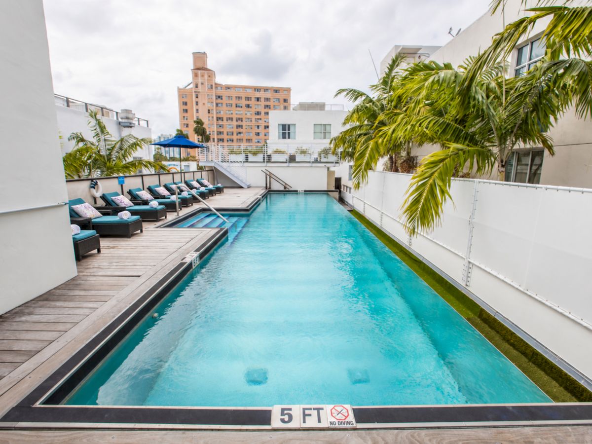 A rooftop swimming pool with turquoise water, sun loungers, and palm trees overlooking city buildings under cloudy skies.